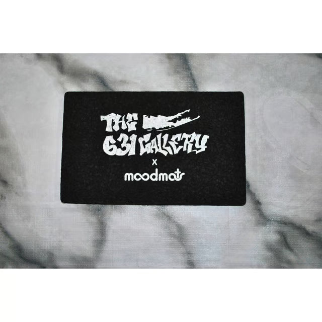 A black rectangular patch featuring white text that reads 'THE 631 GALLERY x moodmat' with a stylized long island graphic, placed on a gray marble-textured surface.