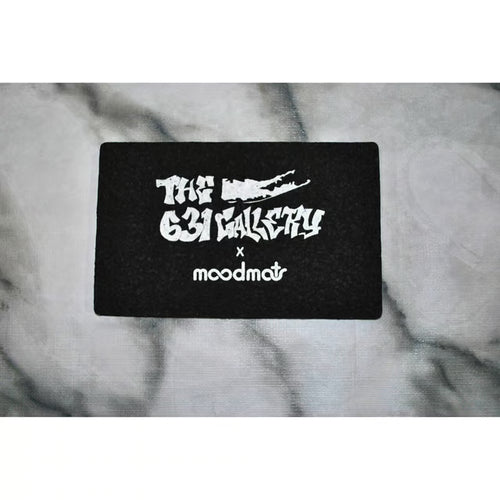 A black rectangular patch featuring white text that reads 'THE 631 GALLERY x moodmat' with a stylized long island graphic, placed on a gray marble-textured surface.