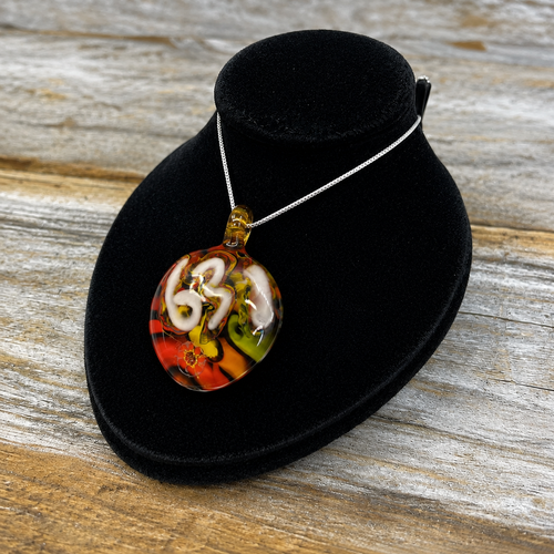 A round glass pendant necklace with a colorful swirl pattern of orange, red, white, and green, displayed on a black velvet jewelry bust over a rustic wooden surface.