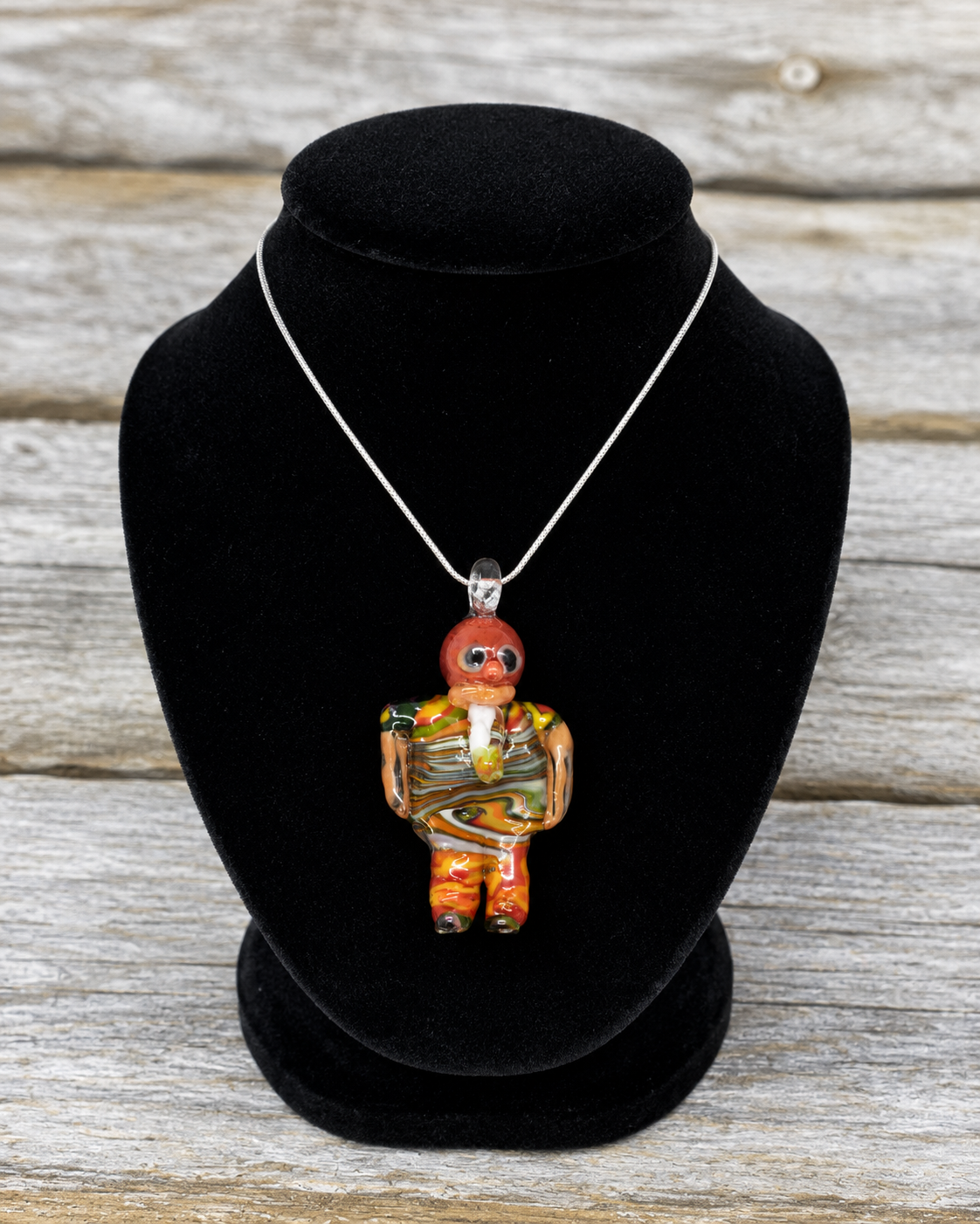 A handmade glass pendant necklace featuring a stylized figure holding a guitar, displayed on a black velvet jewelry bust against a rustic gray wood background.