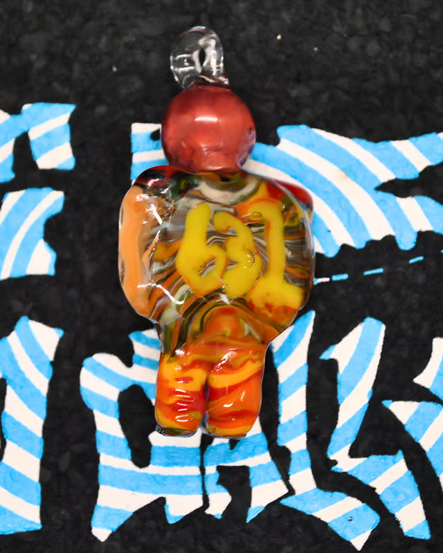 A hand-blown glass pendant shaped like a stylized human figure, featuring a red head, orange and yellow body details, and a clear glass bail at the top, set against a black background with blue and white graphic patterns.