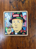 A square cork coaster featuring a colorful cartoon illustration of a man wearing a Supreme hat and holding a cigar, placed on a dark wood tabletop.