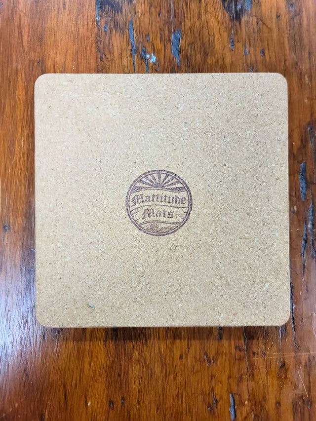 A square tan cork coaster with rounded corners, featuring a circular brown 'Latitude Mats' logo in the center, placed on a dark wood tabletop.