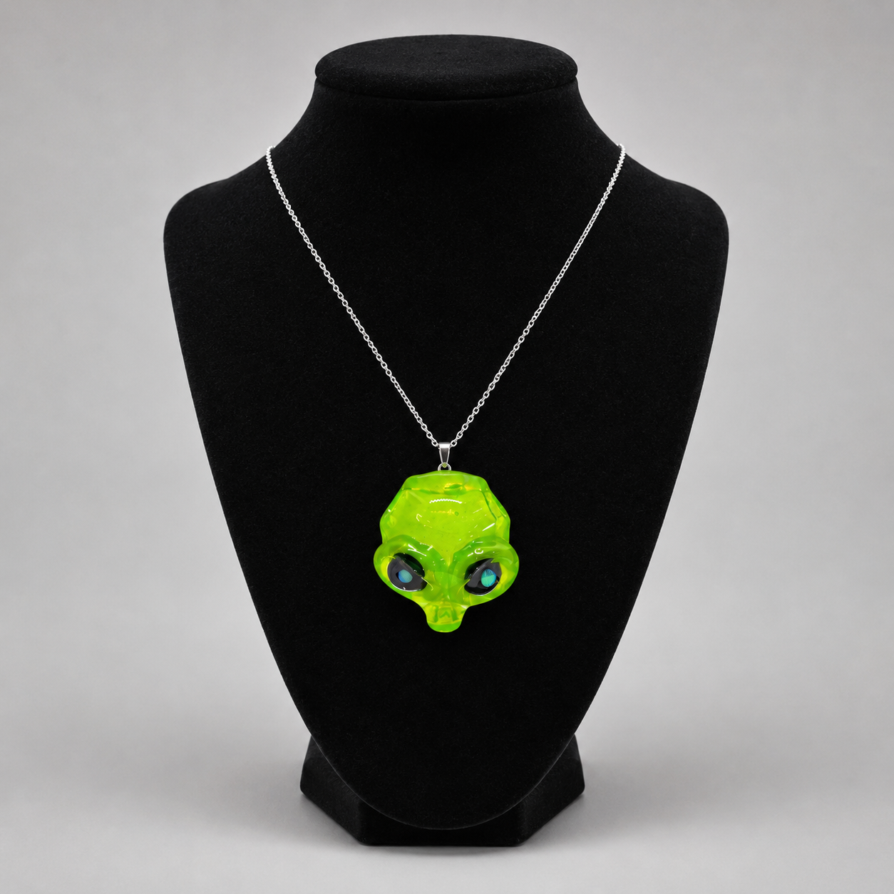A neon green alien head pendant necklace on a silver chain, displayed on a black velvet jewelry bust against a light gray background.