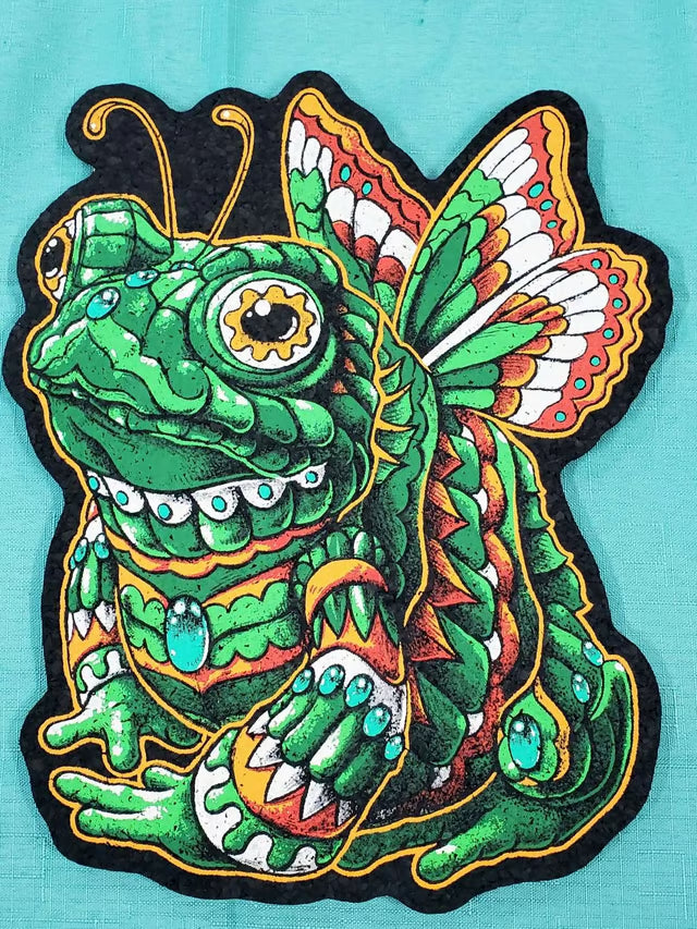 A colorful embroidered patch featuring a stylized green frog-butterfly creature with patterned wings, turquoise accents, and a black outline on a turquoise fabric background.