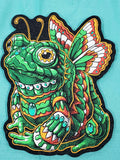 A colorful embroidered patch featuring a stylized green frog-butterfly creature with patterned wings, turquoise accents, and a black outline on a turquoise fabric background.