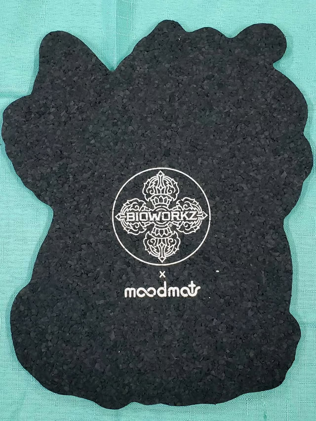 A black textured silicone or rubber accessory with an irregular, organic shape, featuring a white printed logo for 'BIOWORKS x moodmat' in the center, laid flat on a teal background.