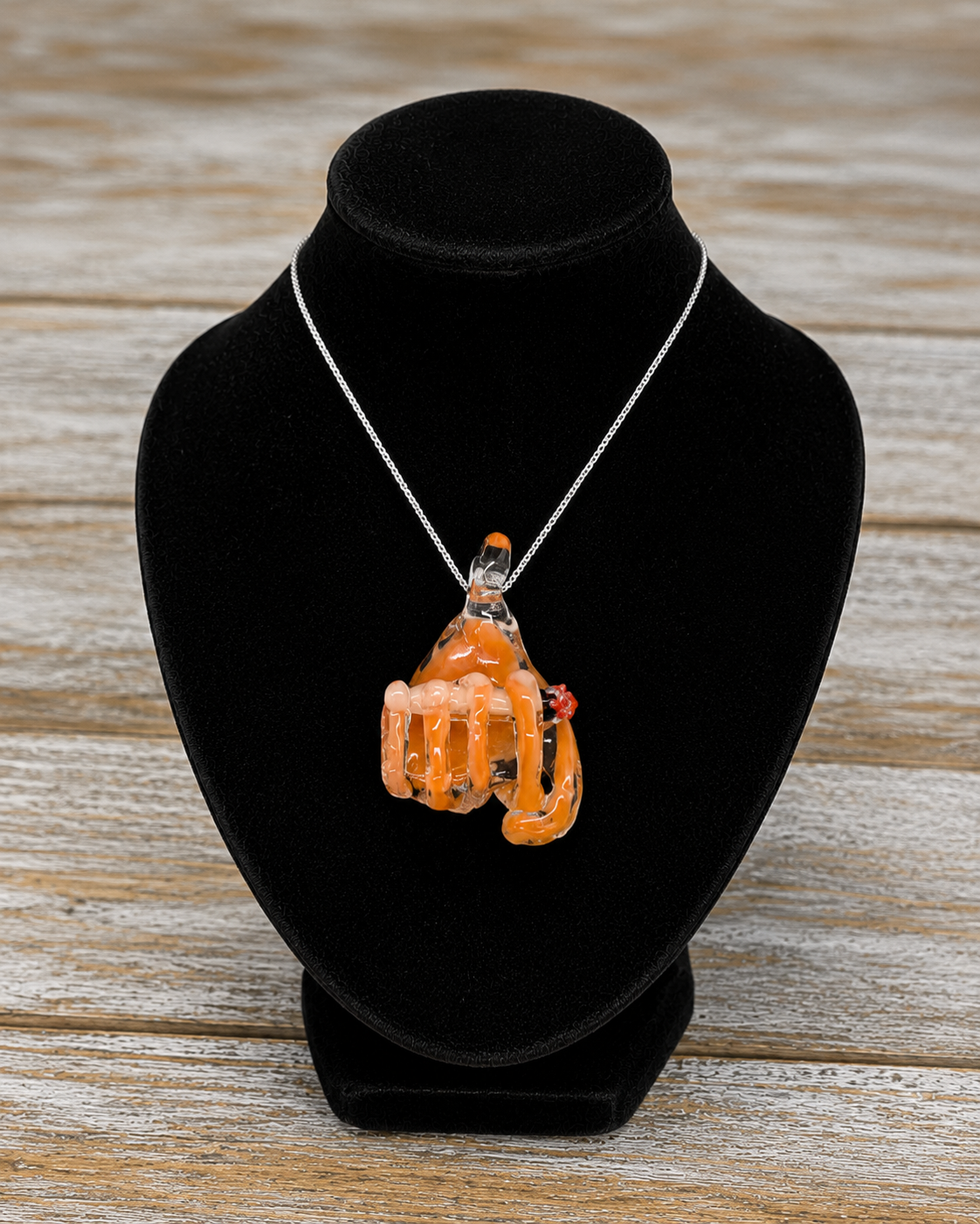 A  glass hand with orange-colored translucent wraps and a small red accent, shown against a velvet jewelry bust against a rustic wooden background.