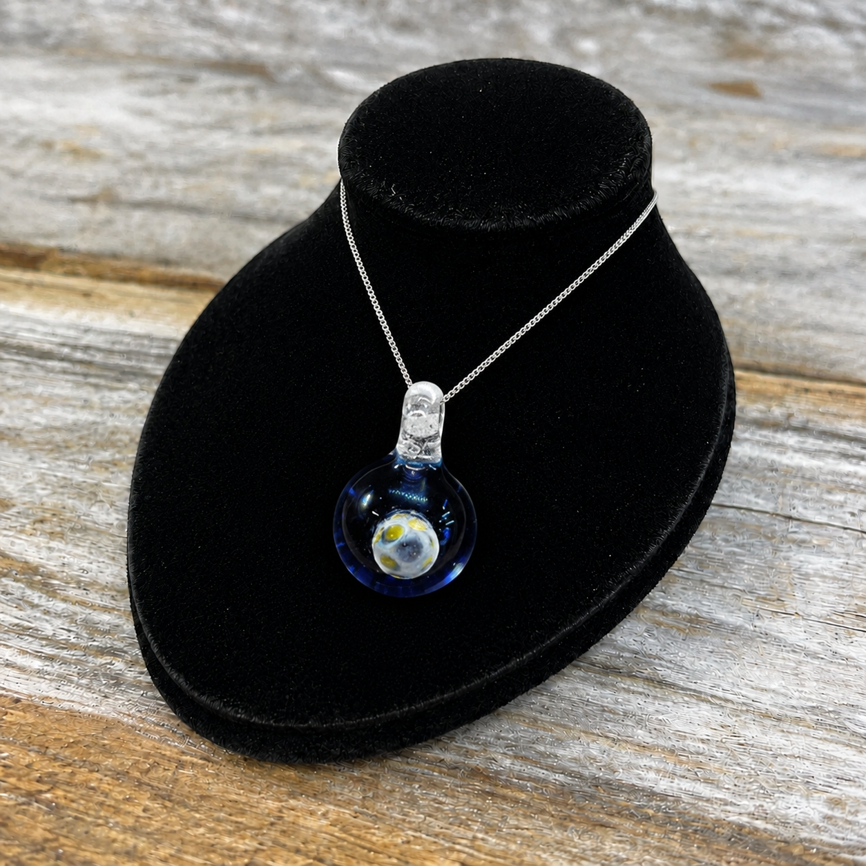 A silver chain necklace with a clear glass bubble pendant containing a miniature blue and white globe, displayed on a black velvet jewelry bust over a rustic wooden surface.