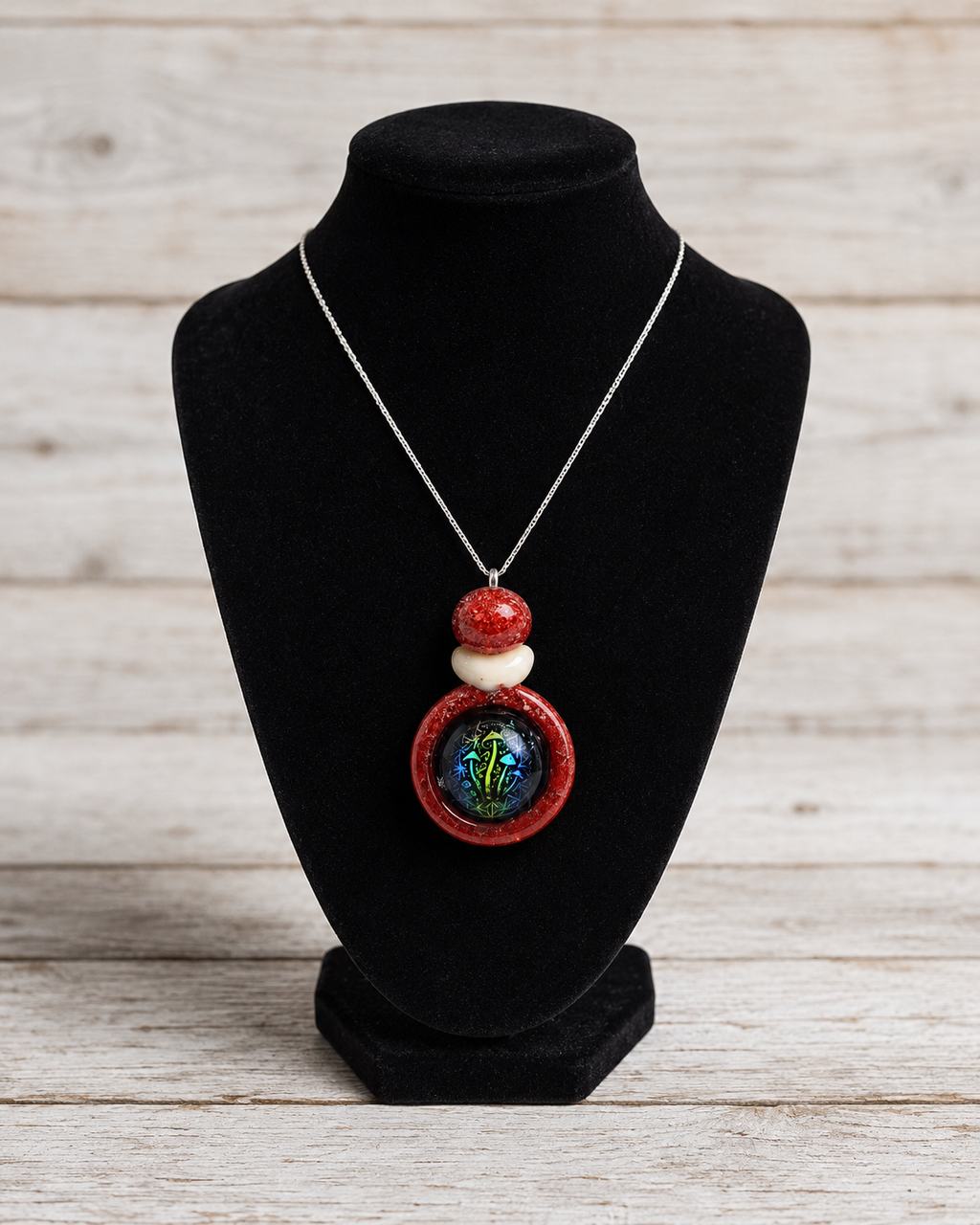 A silver chain necklace with a pendant featuring red and white beads and a round iridescent green-blue center, displayed on a black velvet jewelry bust against a light wood plank background.