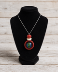 A silver chain necklace with a pendant featuring red and white beads and a round iridescent green-blue center, displayed on a black velvet jewelry bust against a light wood plank background.