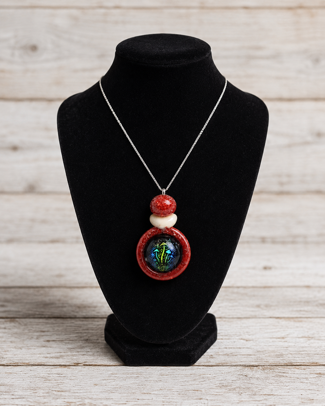 A silver chain necklace with a pendant featuring red and white beads and a round iridescent green-blue center, displayed on a black velvet jewelry bust against a light wood plank background.