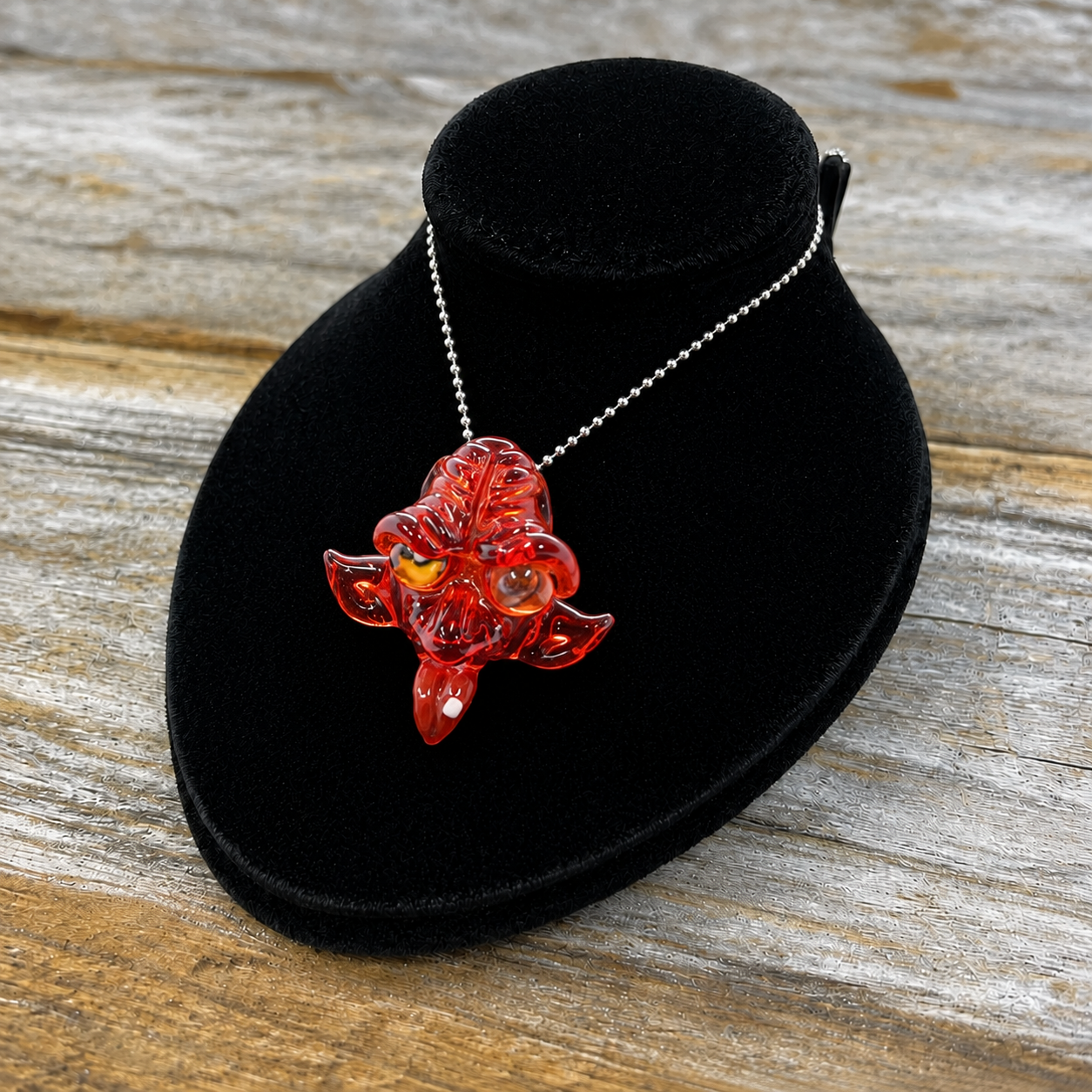 A red translucent dragon pendant necklace on a silver ball chain, displayed on a black velvet jewelry bust over a rustic wooden surface.
