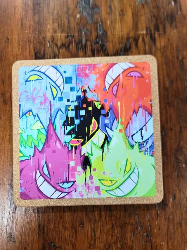 A square cork coaster featuring a vibrant, multicolored graffiti-style graphic of three cartoon monster faces, placed on a dark wooden tabletop.