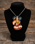 A colorful glass pendant necklace displayed on a black velvet jewelry bust against a rustic wooden background. The pendant features a whimsical character design with blue circular elements, a green patterned center, and orange, red, and white details, suspended from a silver chain.