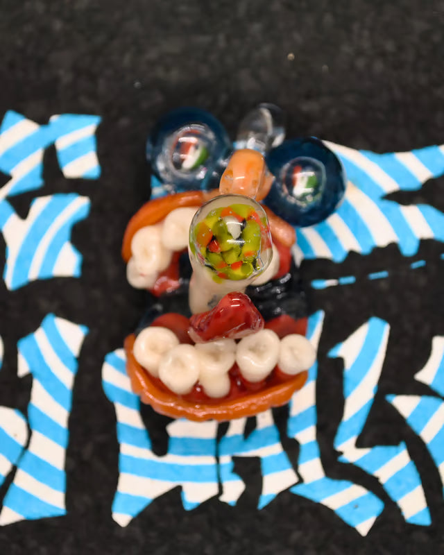 A close-up of a small glass mouthpiece or decorative glass object with white teeth-like forms, orange-red curved glass elements, and a clear central section filled with green, red, and yellow flecks. It is photographed on a black surface with blue-and-white striped paper underneath.