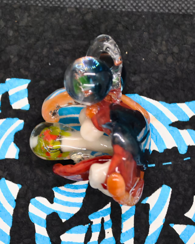 A colorful hand-blown glass ornament or pendant featuring a stylized figure with a clear glass head, red and orange body details, white accents, and dark blue sections, resting on a black textured surface with a blue and white patterned fabric underneath.