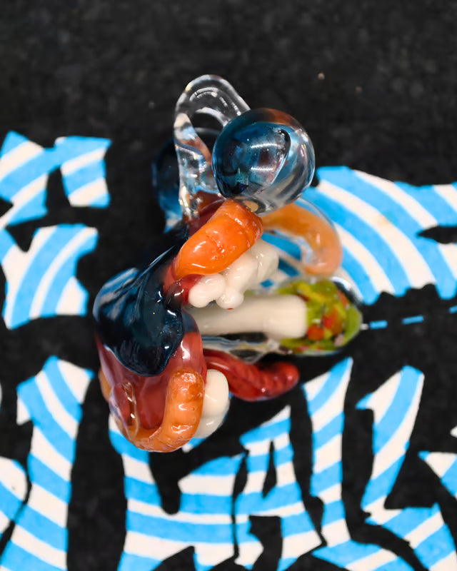 A colorful glass sculpture featuring abstract organic shapes in blue, orange, red, white, and green, set against a black background with a blue and white patterned surface.