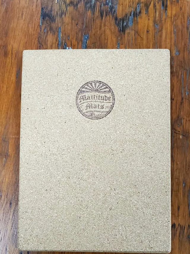 A square cork board or notebook cover in a natural tan color, featuring a circular embossed logo that reads 'Latitude Mats' in the center, placed on a rustic wooden surface.