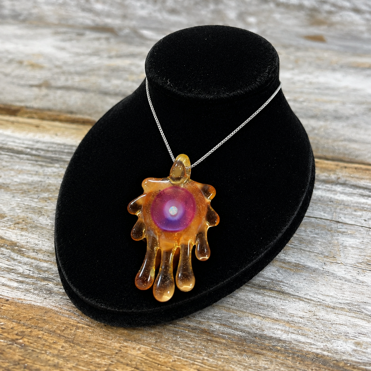 A Glass pendant necklace with a purple and pink center and amber-colored dripping edges, displayed on a black velvet stand over a rustic wooden surface.