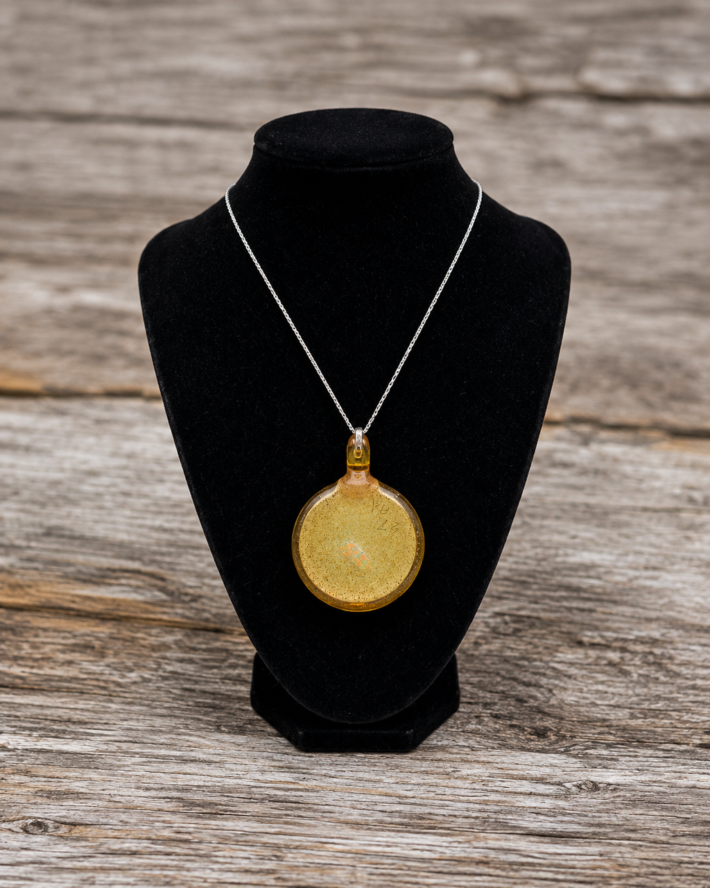 A round gold pendant necklace with a textured finish, displayed on a black velvet jewelry bust against a rustic wooden background.