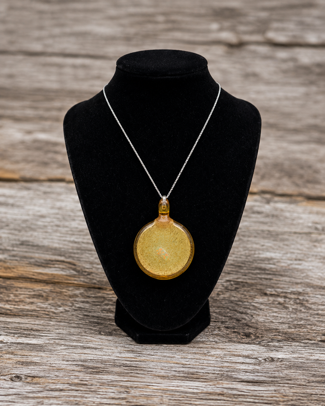 A round gold pendant necklace with a textured finish, displayed on a black velvet jewelry bust against a rustic wooden background.