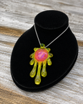 A unique pendant necklace featuring a translucent Lime Green, dripping hand-shaped design with a glowing pink and orange central core, displayed on a black velvet stand over a rustic wood background.