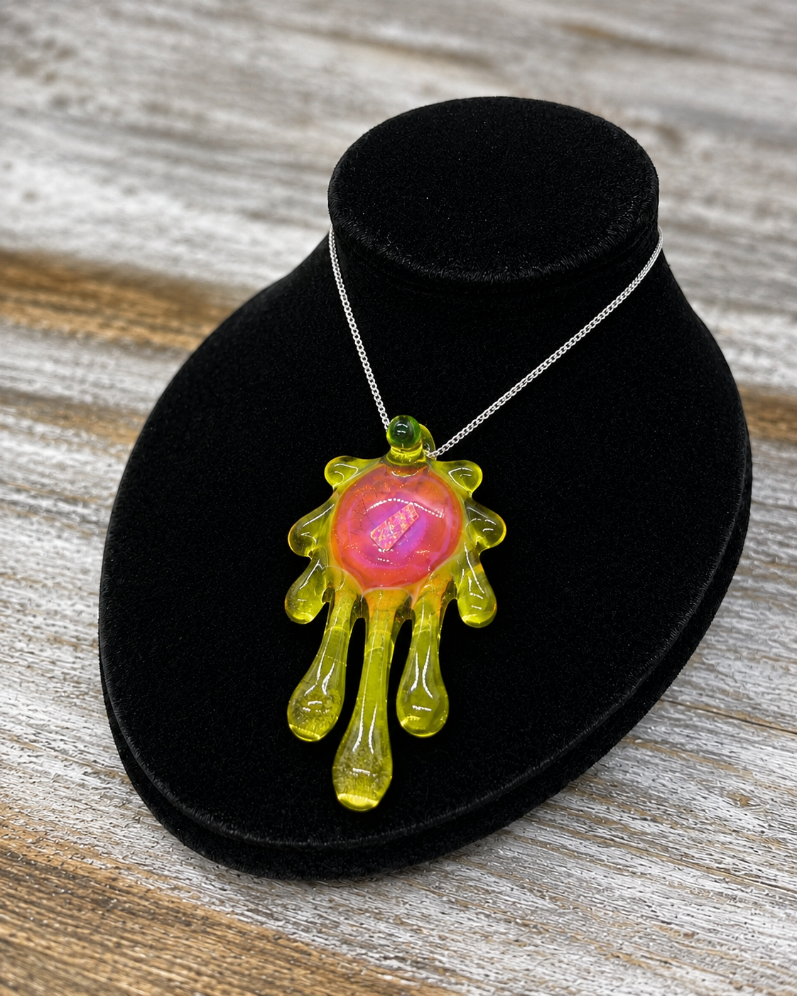 A unique pendant necklace featuring a translucent Lime Green, dripping hand-shaped design with a glowing pink and orange central core, displayed on a black velvet stand over a rustic wood background.