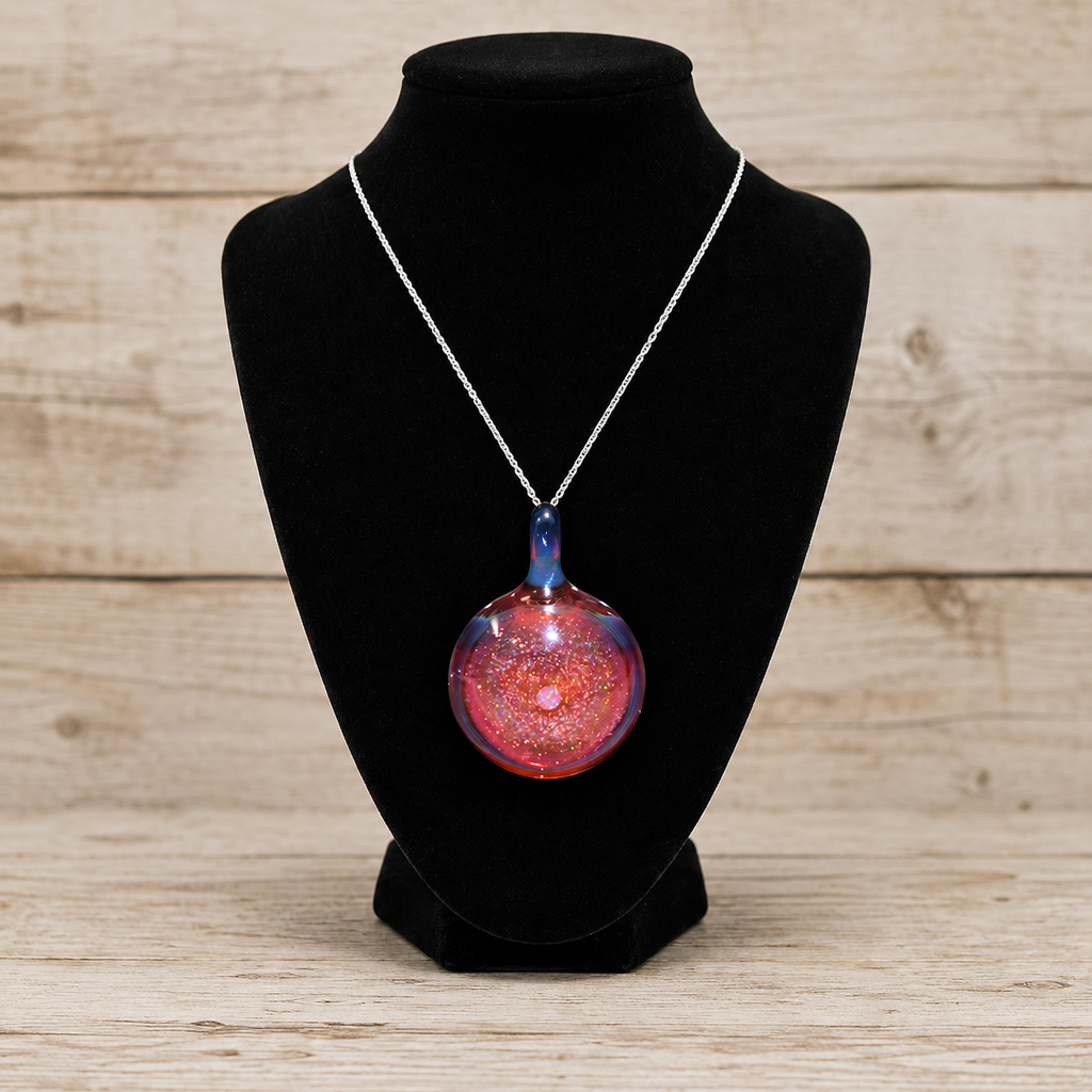 A silver chain necklace with a round glass pendant containing red, pink, and blue glitter, displayed on a black velvet jewelry bust against a light wood plank background.
