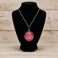 A silver chain necklace with a round glass pendant containing red, pink, and blue glitter, displayed on a black velvet jewelry bust against a light wood plank background.