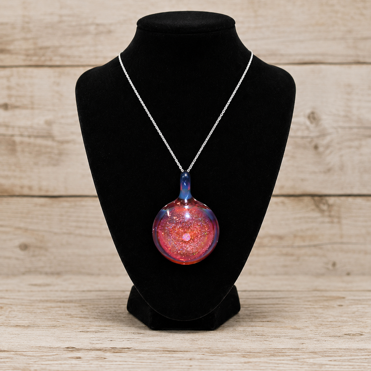 A silver chain necklace with a round glass pendant containing red, pink, and blue glitter, displayed on a black velvet jewelry bust against a light wood plank background.