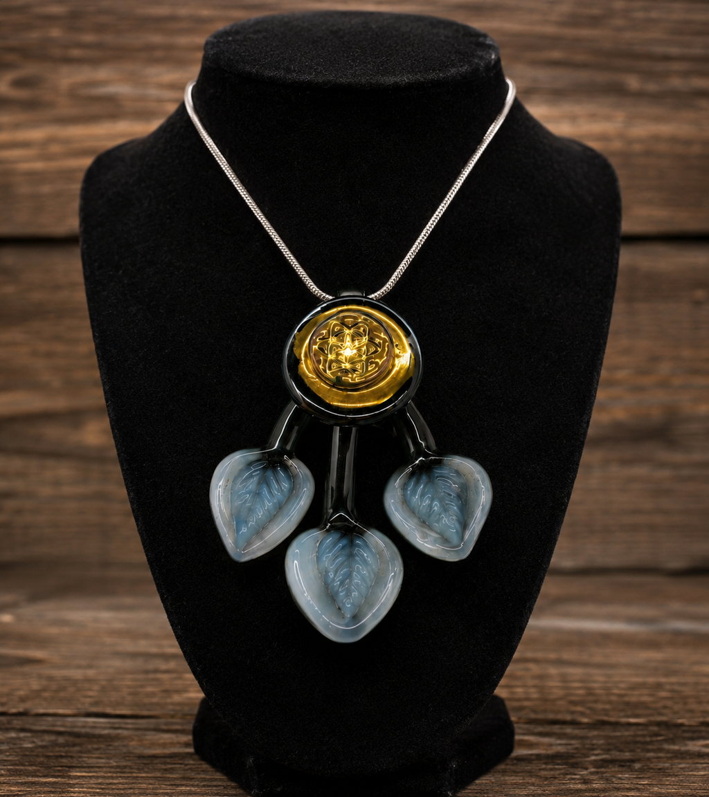 A statement necklace displayed on a black velvet jewelry bust against a rustic wooden background. The necklace features a silver-tone chain, a central round gold-tone medallion with an engraved design, and three dangling translucent light blue glass hearts with a leaf pattern.
