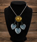 A statement necklace displayed on a black velvet jewelry bust against a rustic wooden background. The necklace features a silver-tone chain, a central round gold-tone medallion with an engraved design, and three dangling translucent light blue glass hearts with a leaf pattern.