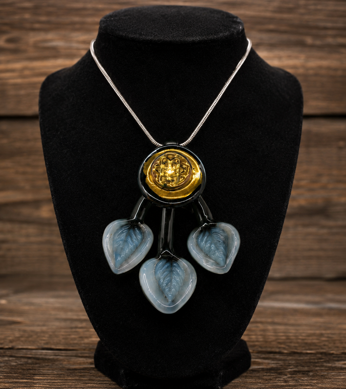A statement necklace displayed on a black velvet jewelry bust against a rustic wooden background. The necklace features a silver-tone chain, a central round gold-tone medallion with an engraved design, and three dangling translucent light blue glass hearts with a leaf pattern.