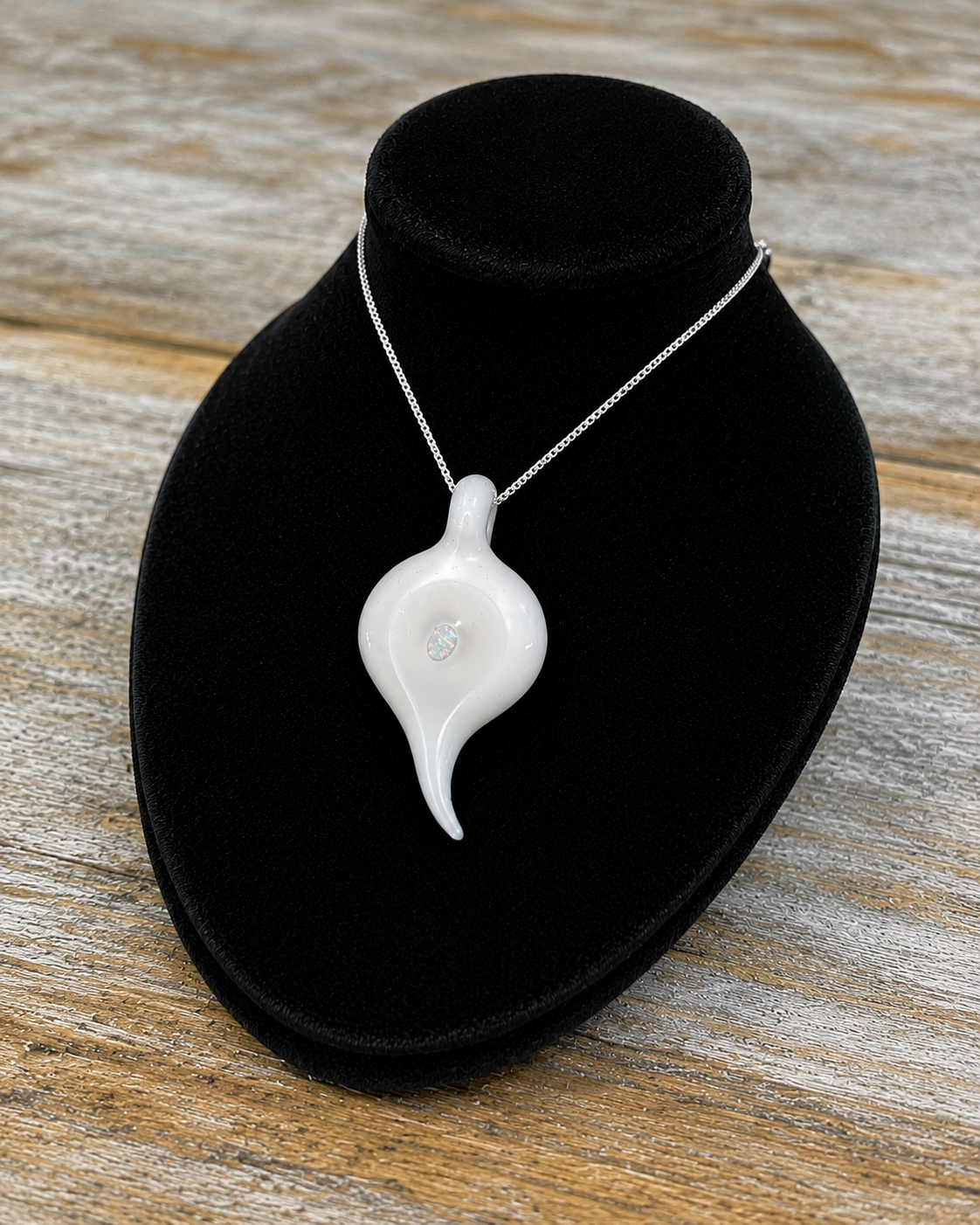 A glossy white Glass teardrop-shaped Pendant with a small loop at the top, resting on a textured black surface on a bust