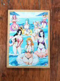 A rectangular printed art piece or book cover featuring colorful anime-style illustrated women in swimwear on a beach, photographed from above on a dark wooden surface.