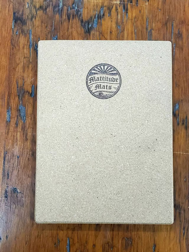 A square beige textured notebook or journal with a circular black 'Latitude Mats' logo on the front, placed on a rustic wooden table.