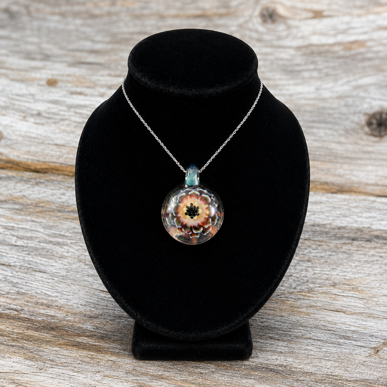 A silver chain necklace with a round glass pendant featuring a colorful floral design, displayed on a black velvet jewelry bust over a rustic wooden surface.