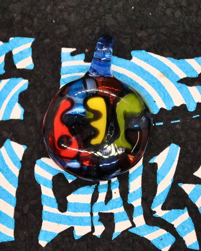 A round glass pendant with a blue loop, featuring swirls of red, yellow, green, and black, placed on a black background with blue and white striped graffiti-style paint.