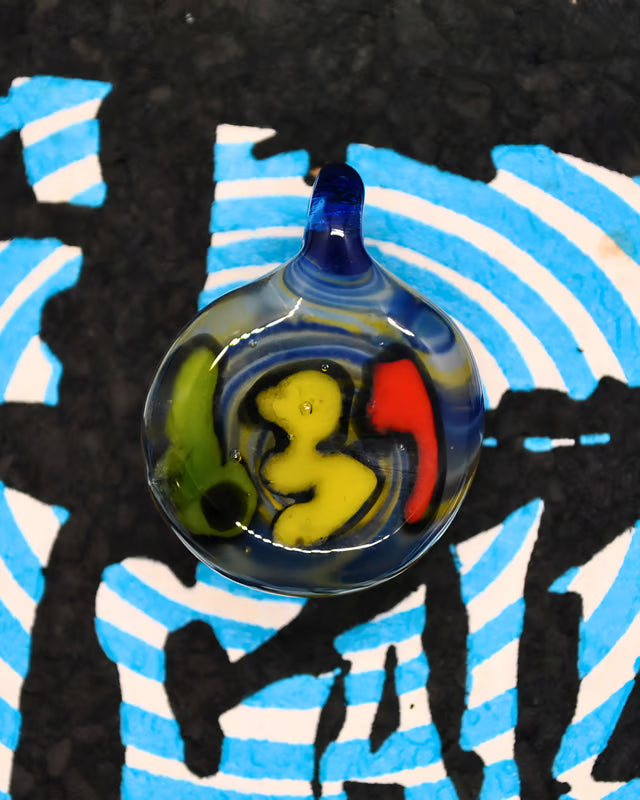 A round glass pendant with a blue stem, featuring a colorful swirl design of yellow, red, green, and blue, resting on a black and blue patterned surface.