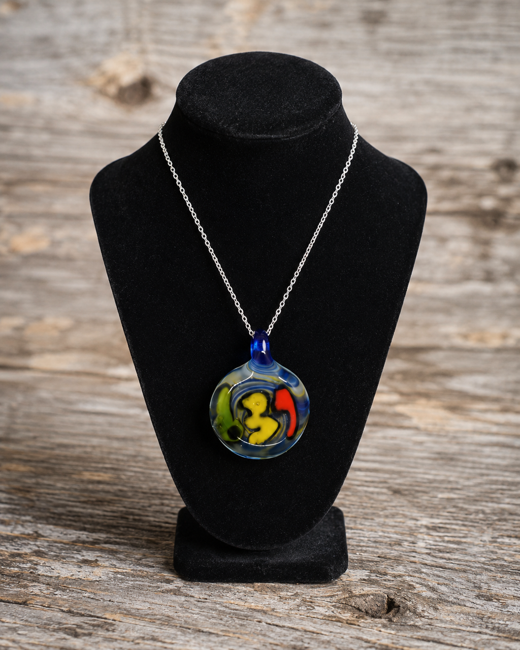 A colorful round pendant necklace displayed on a black velvet jewelry bust against a rustic wooden background. The pendant features abstract swirls of yellow, green, red, and blue glass, suspended from a silver chain.