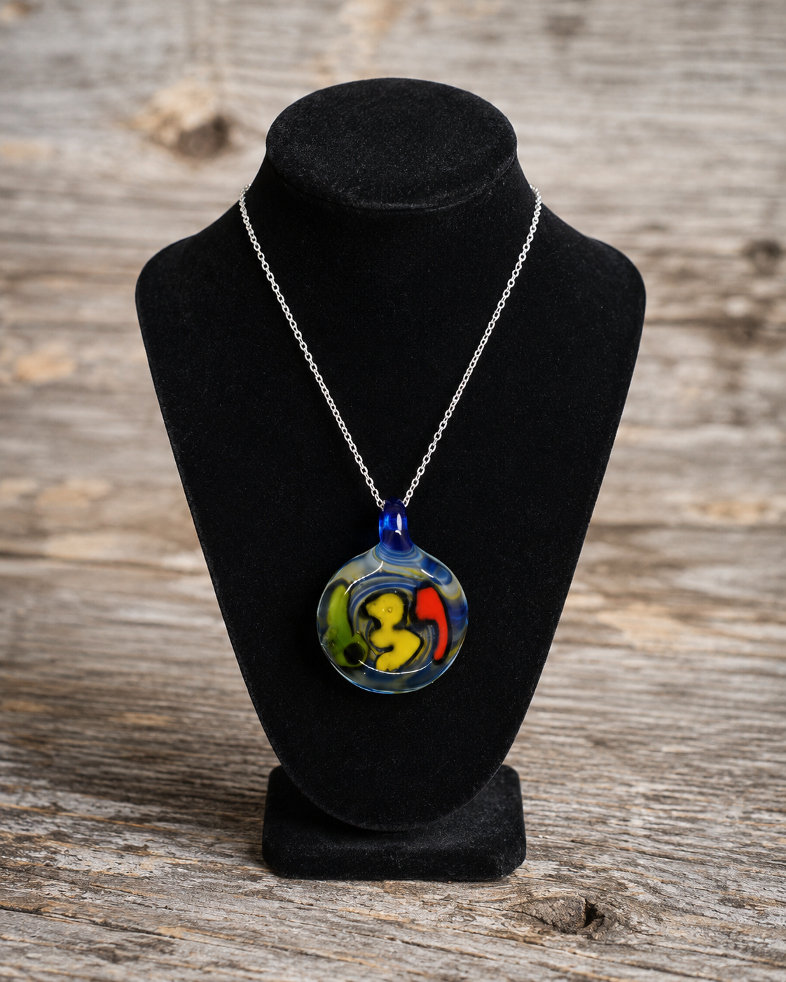 A colorful round pendant necklace displayed on a black velvet jewelry bust against a rustic wooden background. The pendant features abstract swirls of yellow, green, red, and blue glass, suspended from a silver chain.