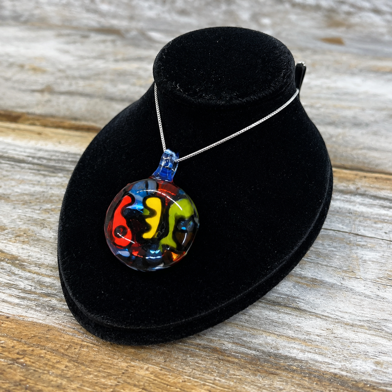 A colorful round glass pendant necklace with a silver chain, displayed on a black velvet jewelry bust over a rustic wooden surface. The pendant features a vibrant, abstract swirl pattern of red, yellow, green, and blue glass.