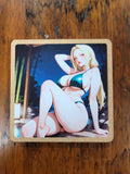 A square printed coaster or tile with rounded corners, featuring an anime-style illustration of a blonde woman in a dark teal bikini sitting at night, placed on a brown wooden surface.