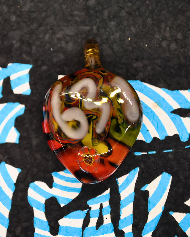 A hand-blown glass pendant with a teardrop shape, featuring swirls of red, orange, yellow, white, and black, resting on a black textured surface with blue and white graphic elements.